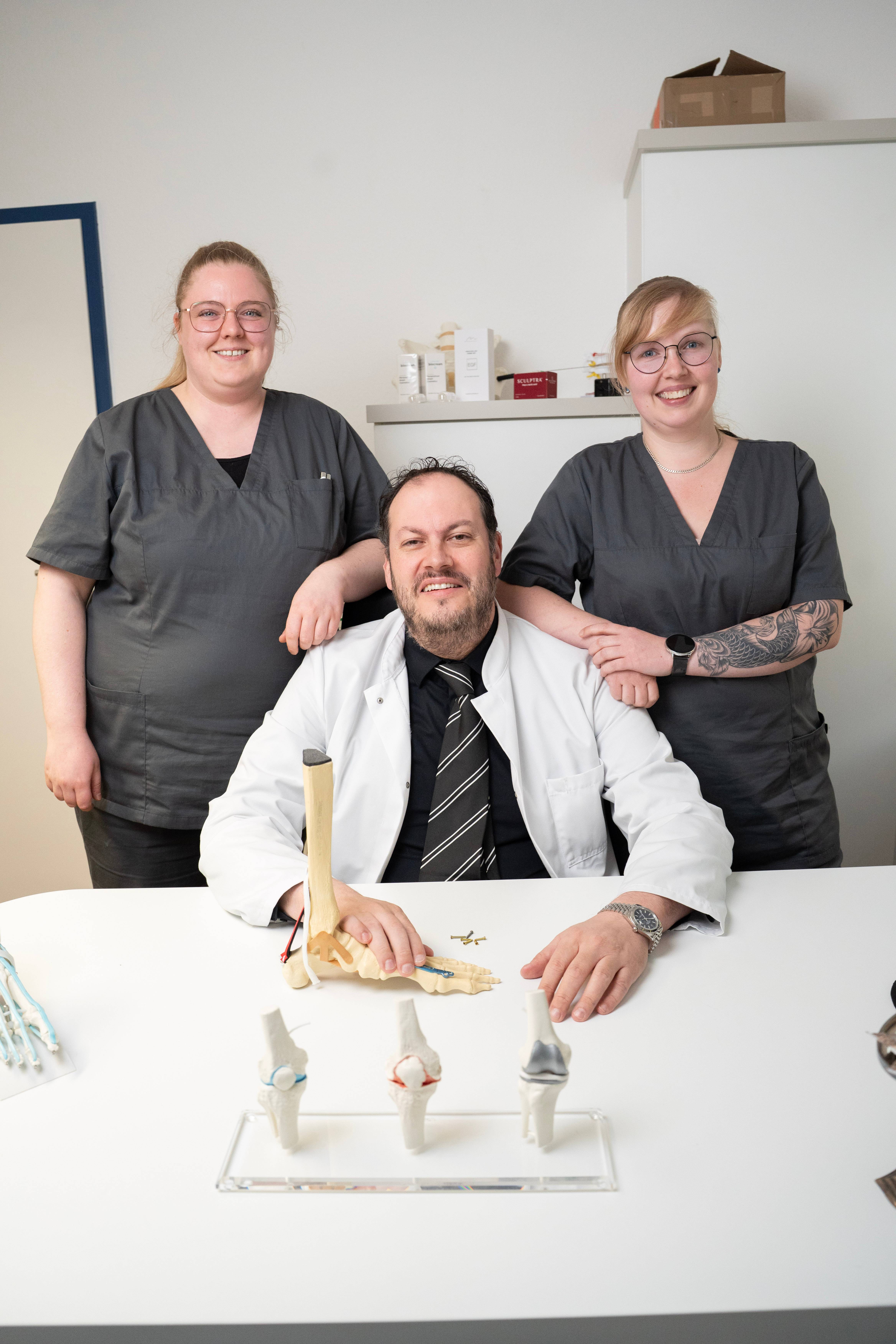 Dr. Velickovic mit zwei Medizinischen Fachangestellten am Schreibtisch mit Knie- und Fußmodellen in der Praxis Gifhorn